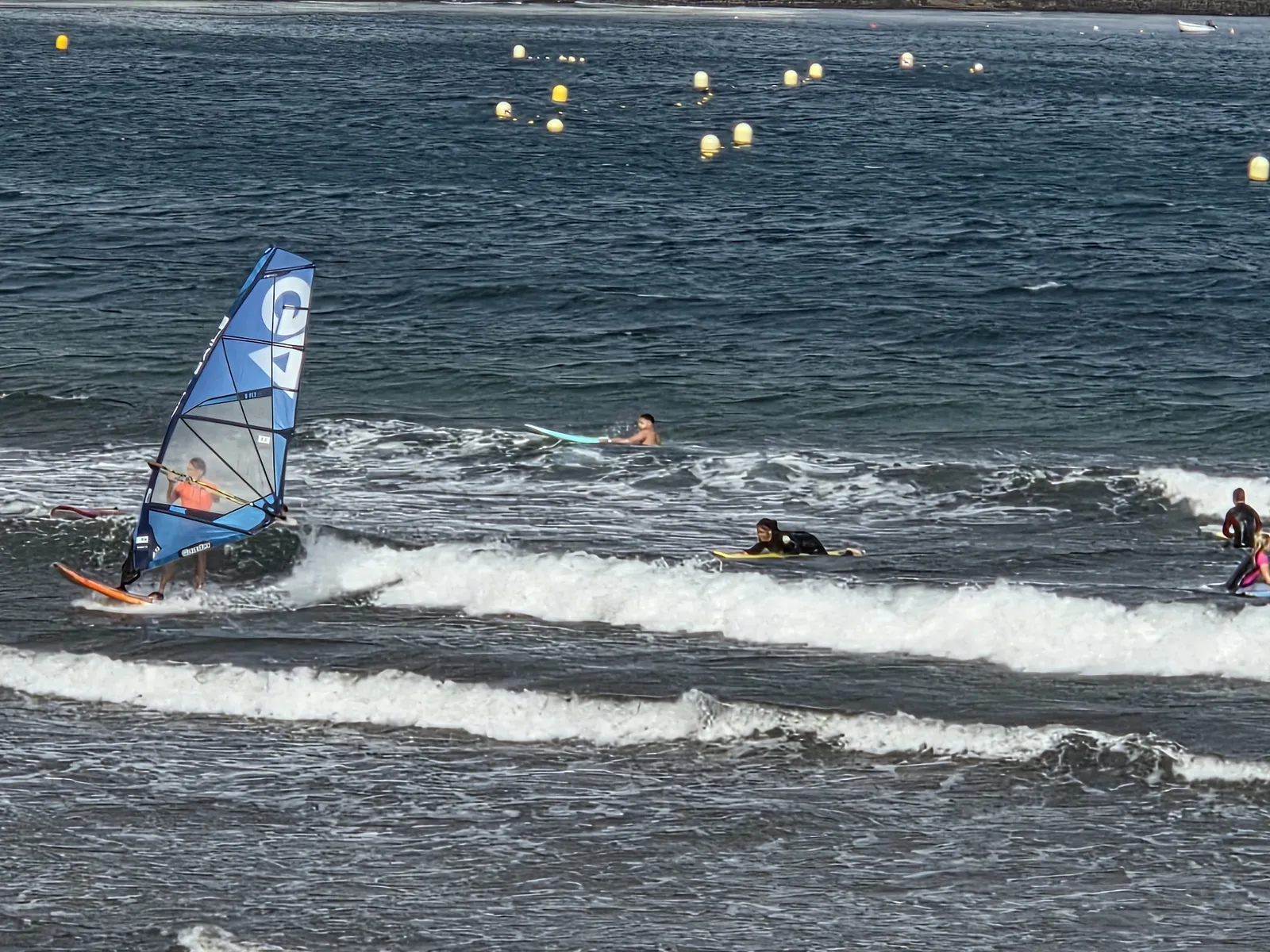 Windsurfer riding a small breaking wave near people on surfboards and bodyboards in El Médano