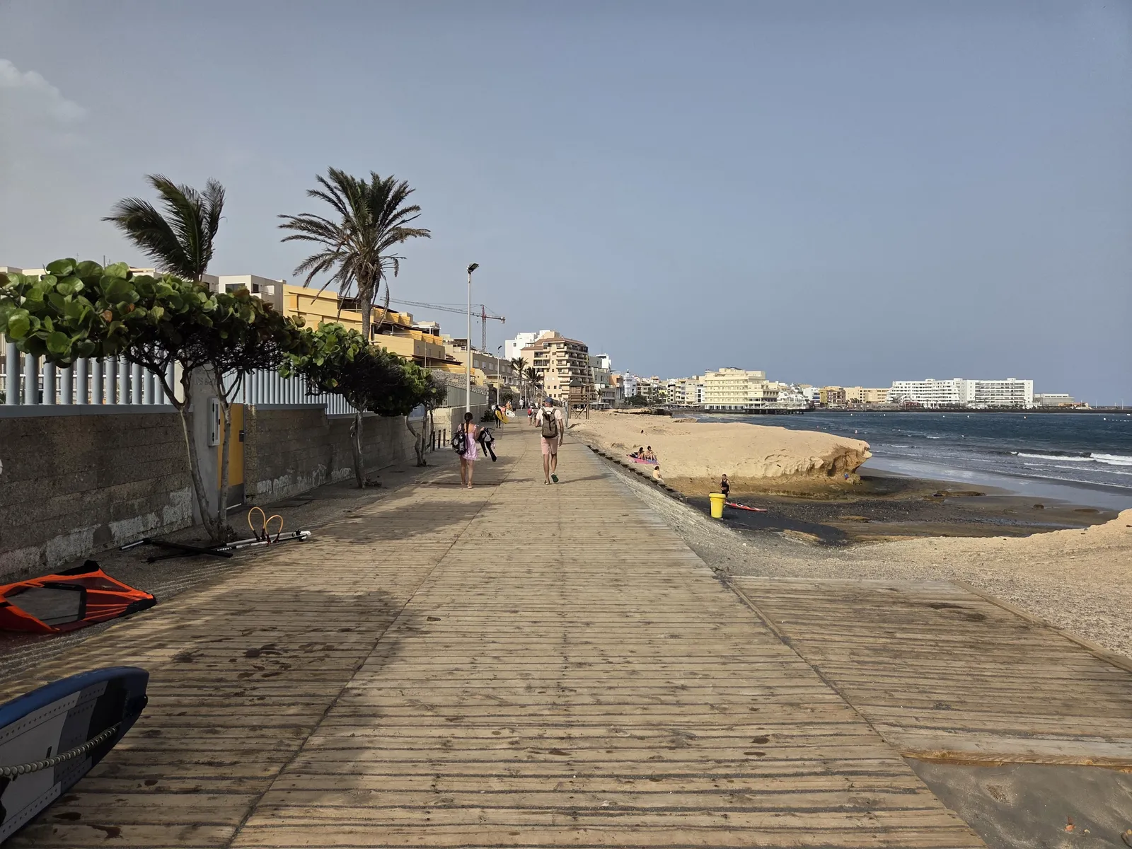 Wooden seafront promenade beside sandy rock formations, with El Médano buildings across the bay