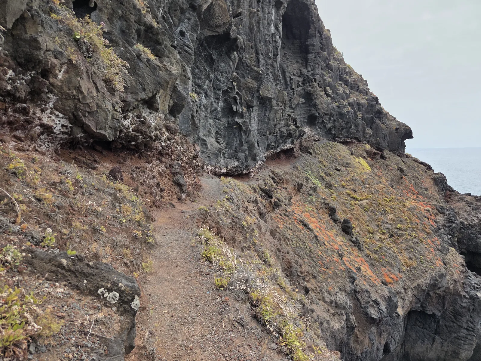 Narrow dirt path beside a rugged coastal cliff, with a steep drop towards the sea
