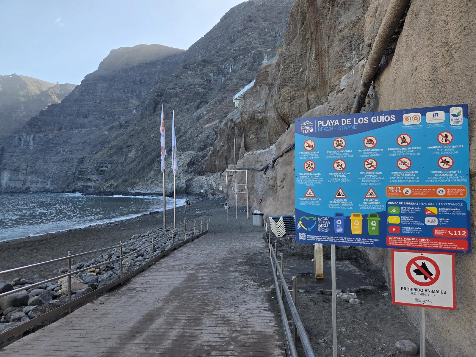 Acceso a Playa de Los Guíos con panel de normas, arena oscura y acantilados junto al mar