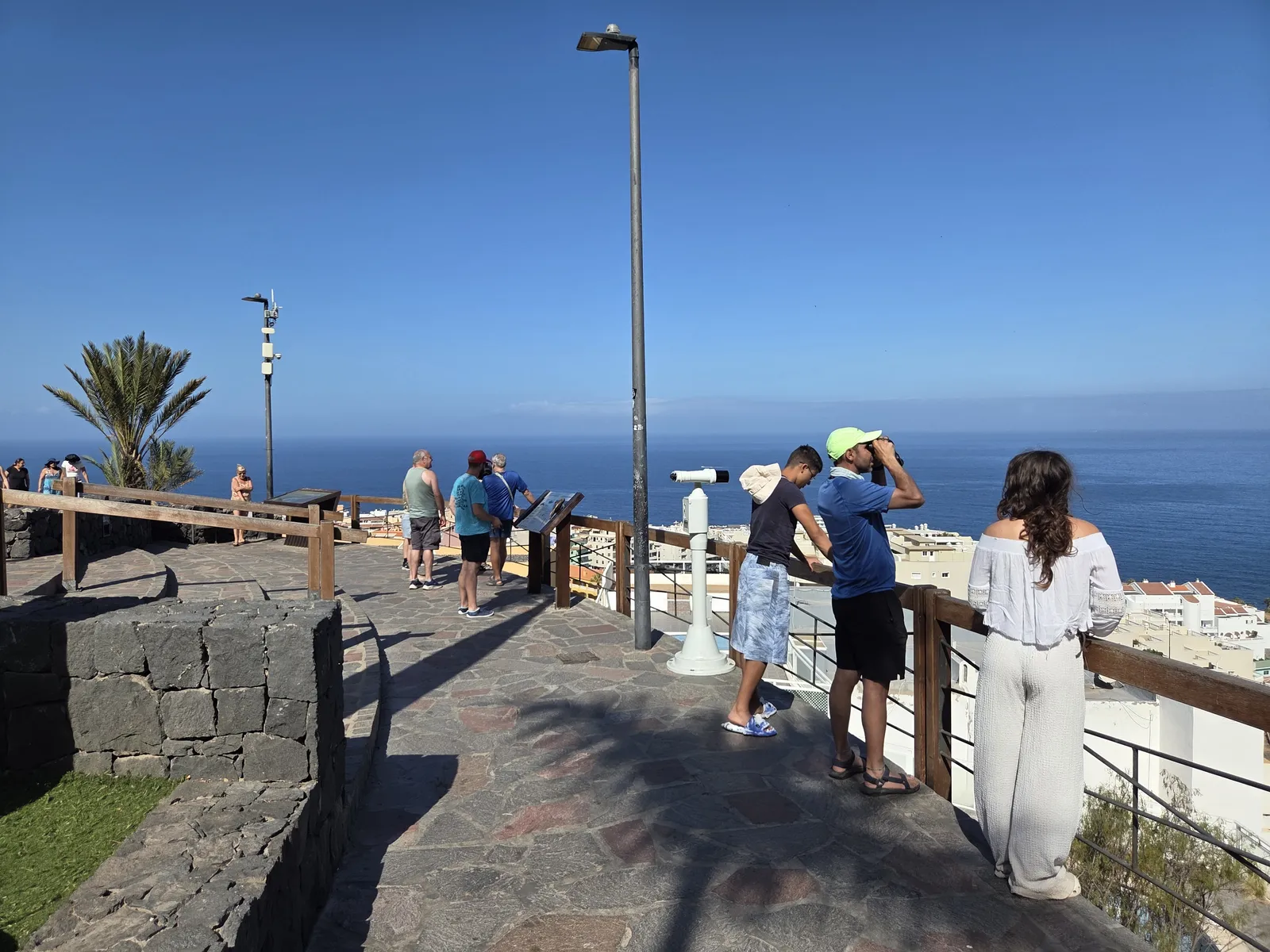 Visitantes junto a la barandilla de un mirador de Los Gigantes, con el océano y edificios costeros al fondo