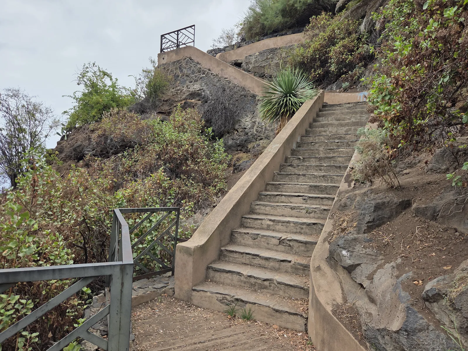 Worn stone steps zigzagging uphill between low walls, coastal vegetation and metal railings