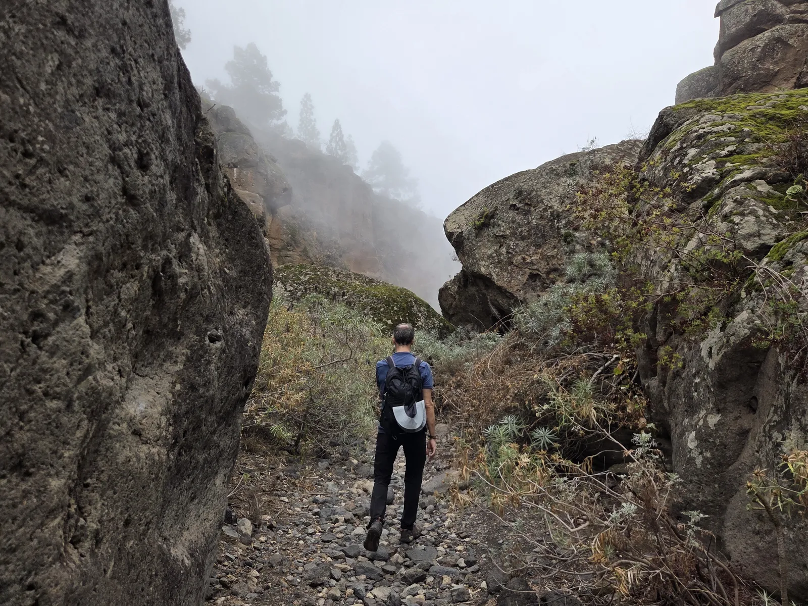 Hiker carrying a backpack along a rocky path between large boulders in mist