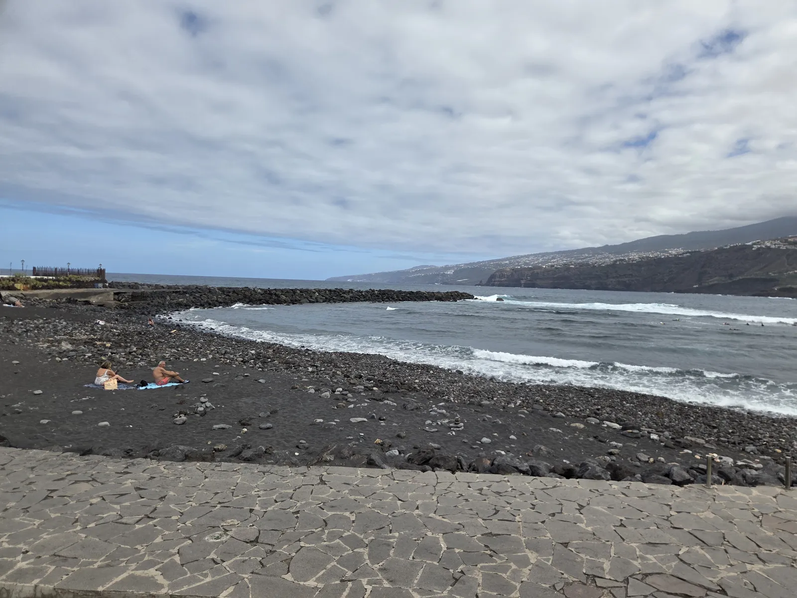 Dark sand and rounded stones at Playa Martiánez, with breaking waves and coastal cliffs beyond