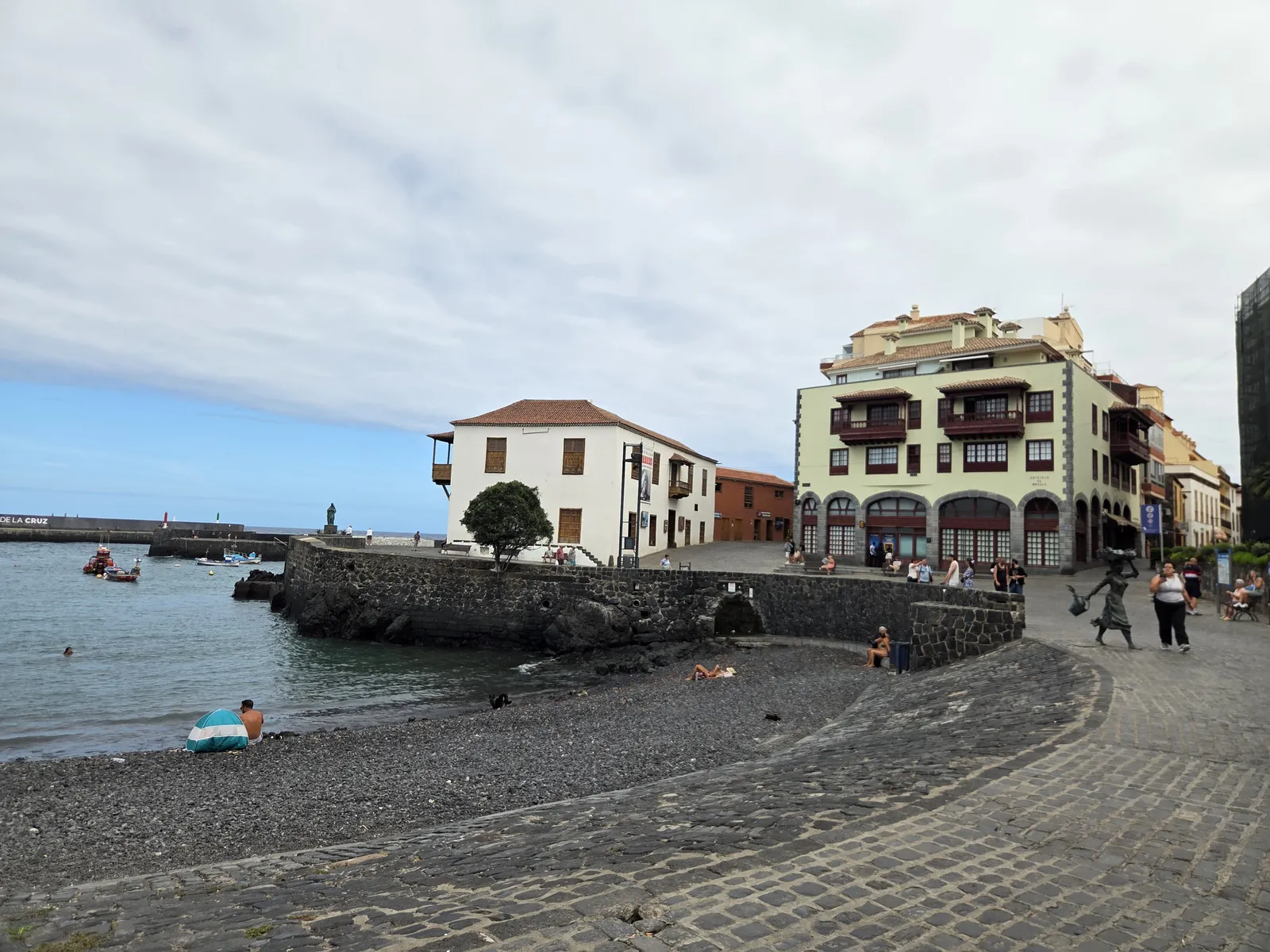 Small dark-pebble beach beside Puerto de la Cruz harbour, with fishing boats and traditional waterfront buildings
